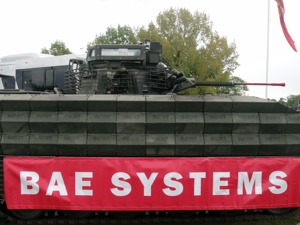 A military tank shown close up with the words 'inert' printed multiple times along its body. A red banner on the tank reads 'BAE SYSTEMS' – the weapons company contracted for police data plans