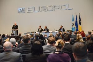 Three people in suits sit behind a panel table in a large space in front of a seated audience. Silver letters read 'Europol' on the wall above, and three flags hand next to the table, including the EU flag and Europol flag.