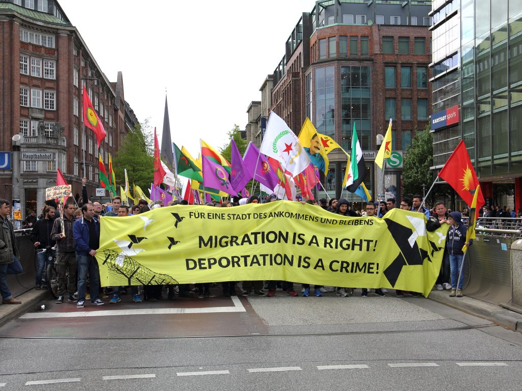 Protesters hold flags of different nations and organisations in a street in Hamburg, holding a large yellow banner reading "Migration is a right! Deportation is a crime!"