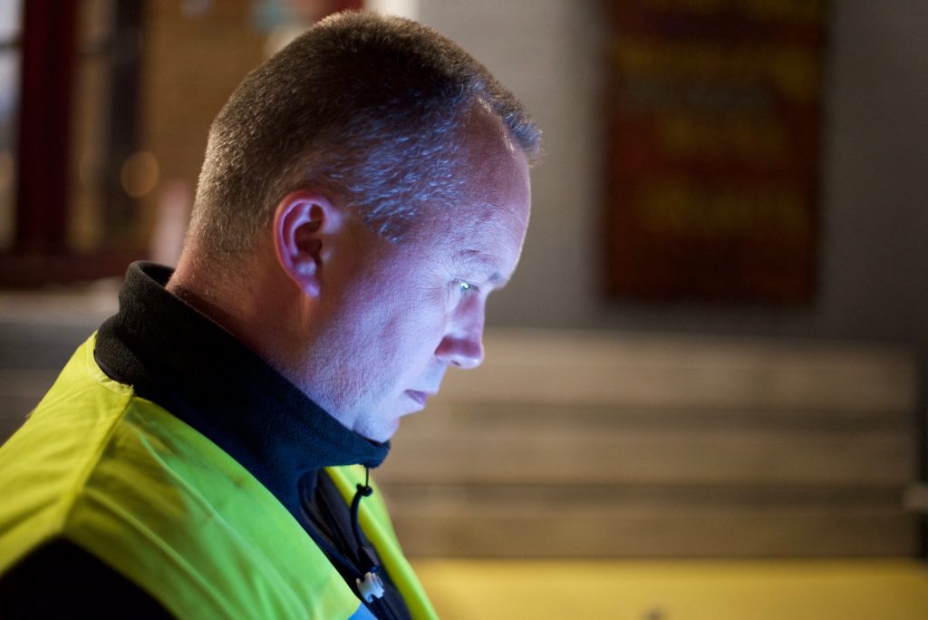 Tomey Olsen, an activist monitoring refugee arrivals in Greece, wears a high-vis yellow jacket. He looks down, his face in profile and illuminated by a screen. Stairs are visible in the shot behind him